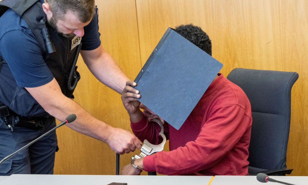 A court official (L), removes the handcuffs from the defendant, who is covering his face with a file folder, in the hearing room of the Regional Court in Ulm, Germany, on July 4, 2023. (Stefan Puchner/dpa via AP)